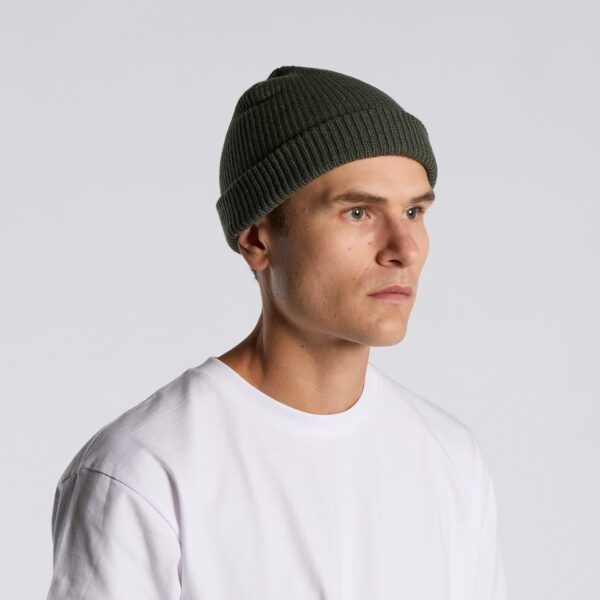 A green ribbed beanie worn by a model with short hair, paired with a white t-shirt. The beanie features a logo.